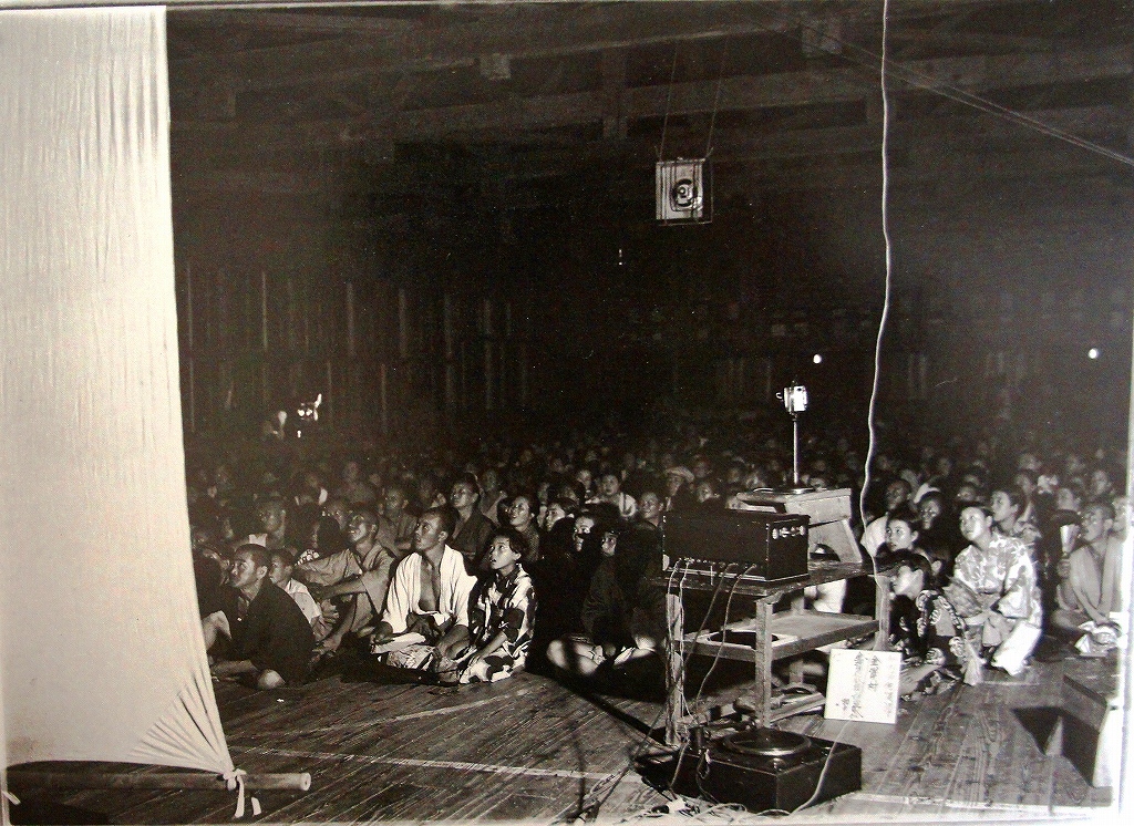 「佐渡万華鏡」 91　昭和初期　金沢村の集会所　活動写真には金沢信販組合員たちが